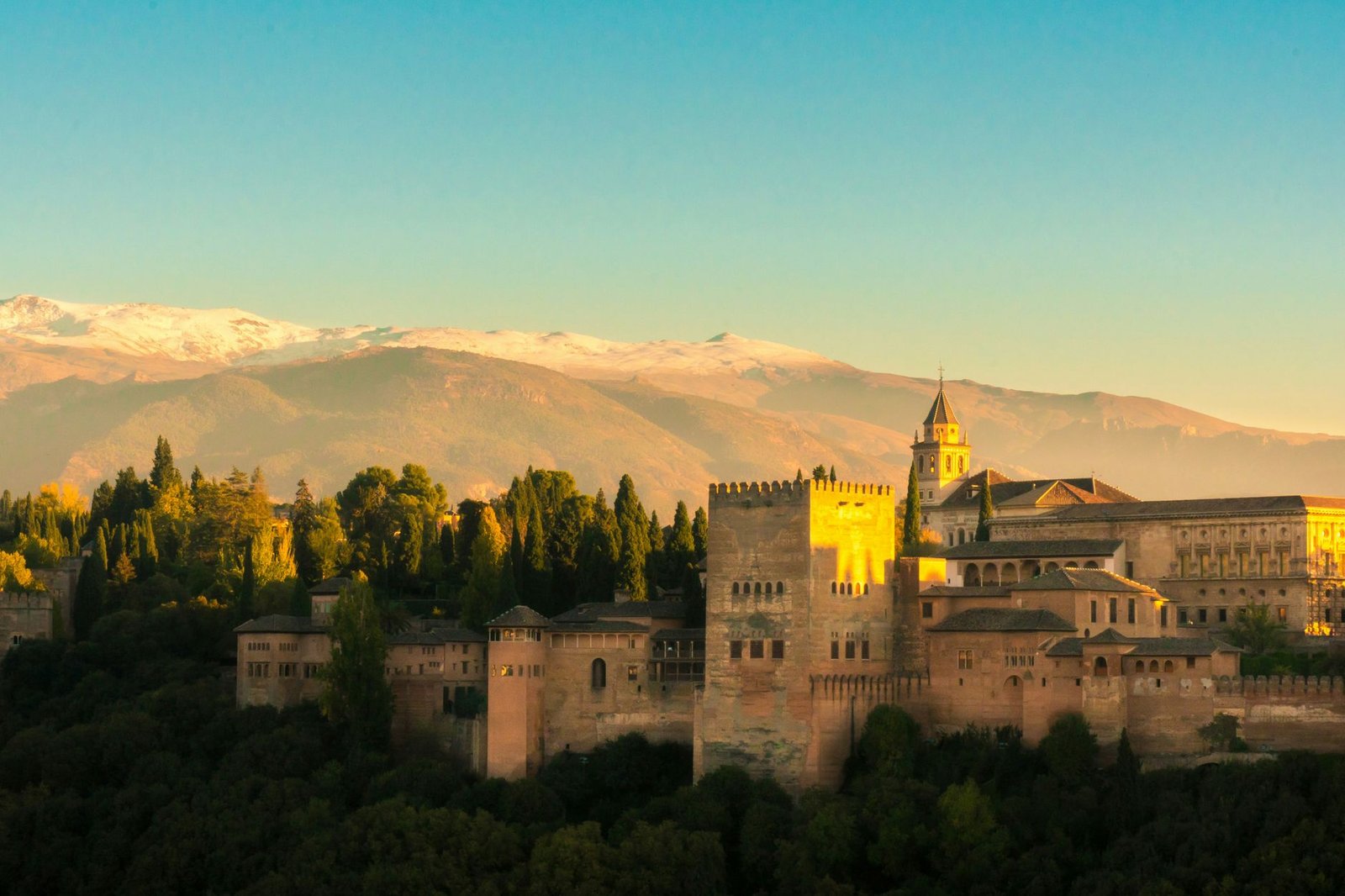 View of Alhambra Palace surrounded by trees with Sierra Nevada mountains in the background at sunset