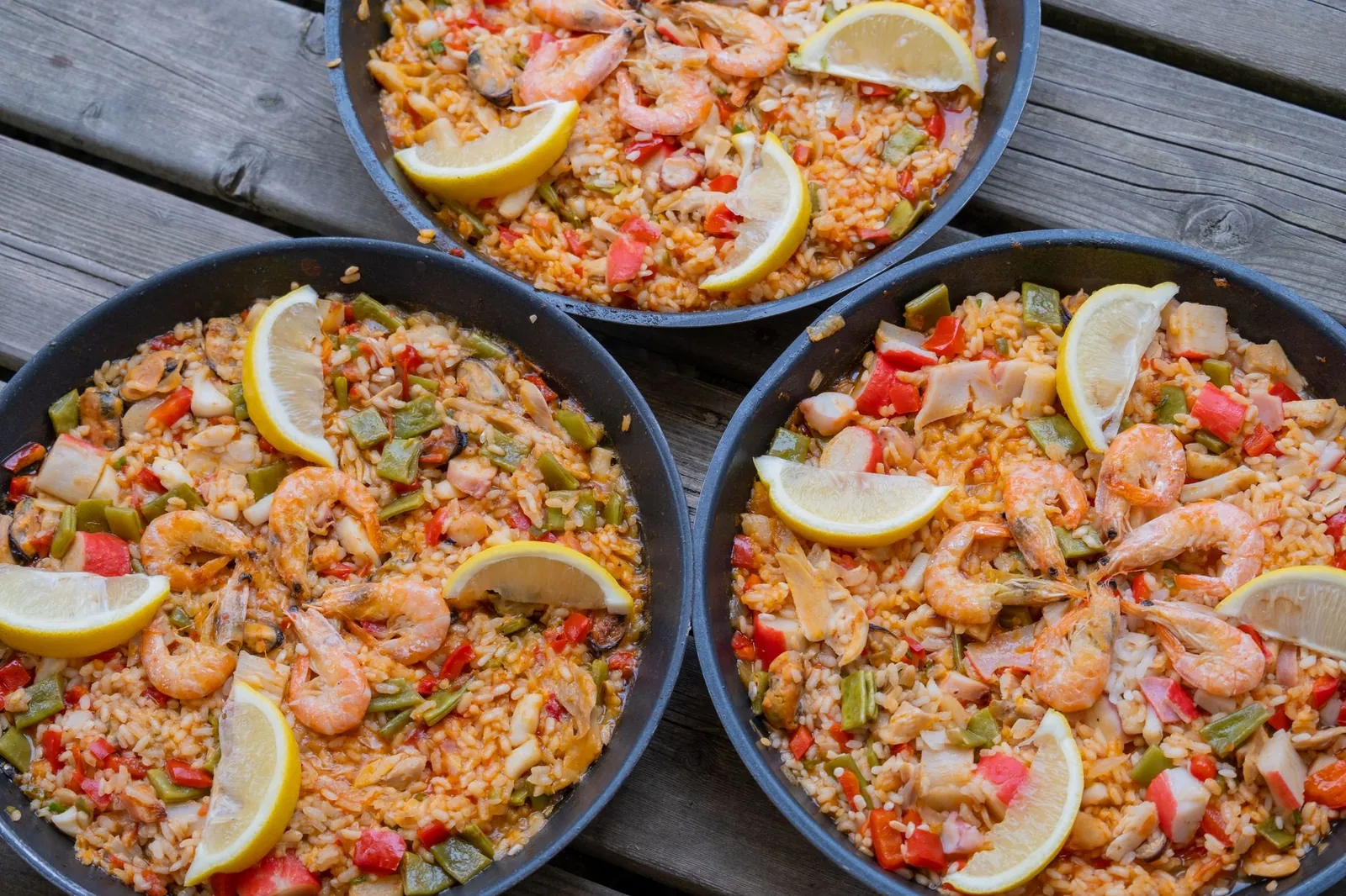 Close-up of three pans of seafood paella with lemon slices on a wooden table