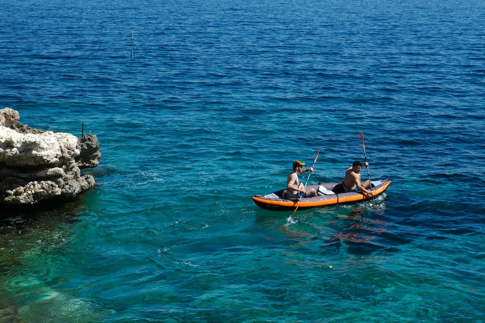 Two men enjoy kayaking in the crystal-clear sea near a rocky shore under the summer sun