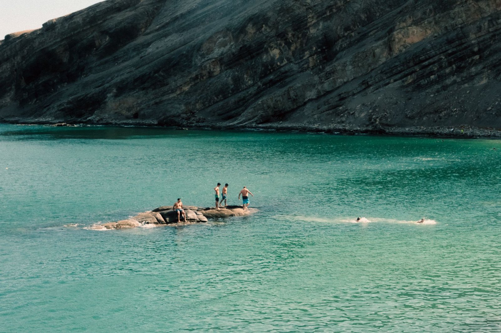 A group of adults swimming and relaxing on rocks in clear turquoise water by rugged cliffs