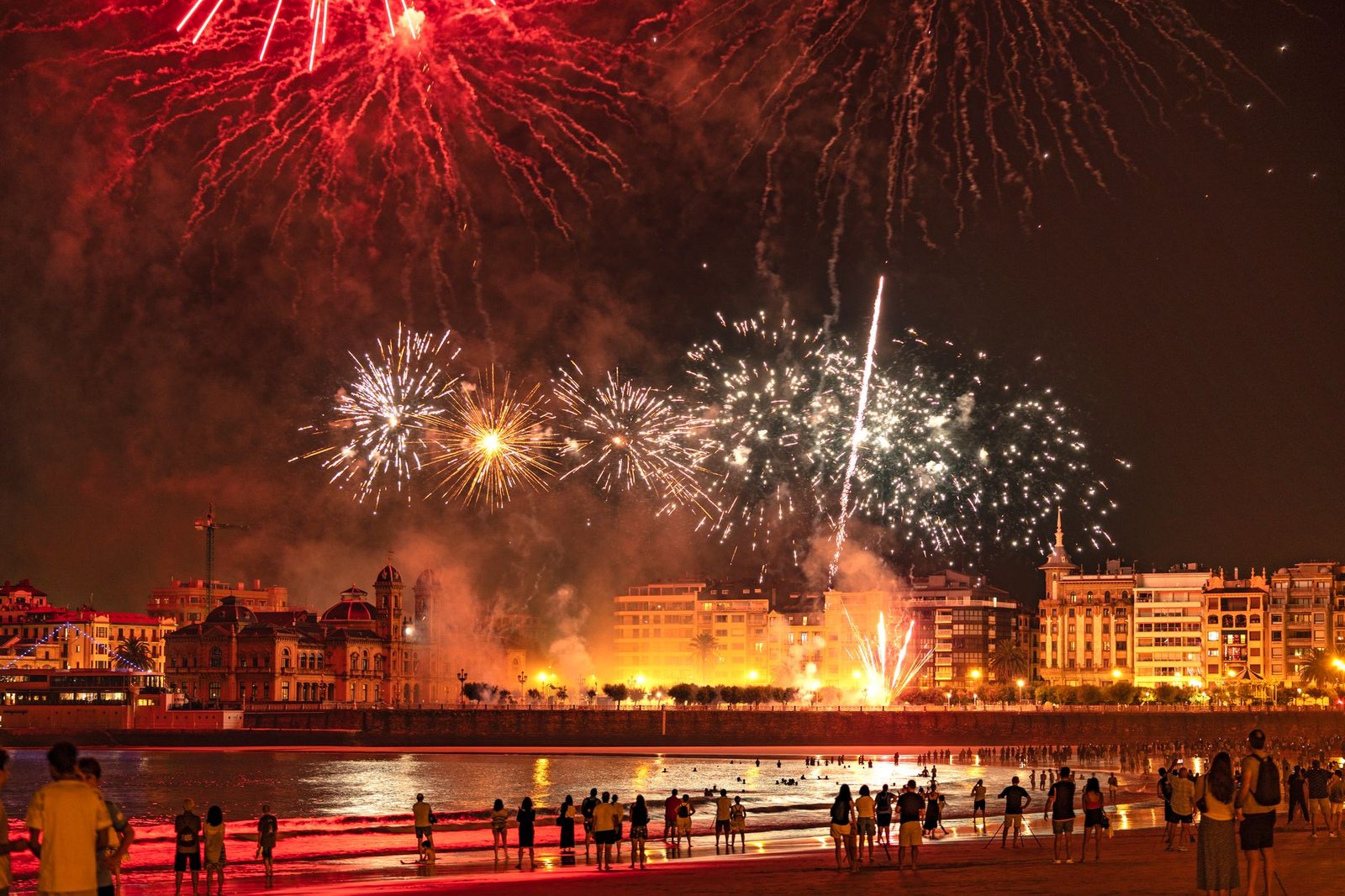 Bright fireworks illuminate a coastal cityscape at night, drawing an audience along the beach