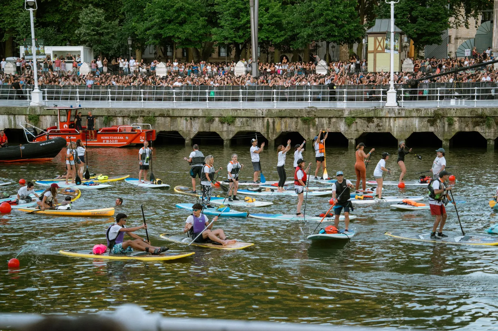 A lively paddleboarding event on a river with a large crowd watching from the embankment