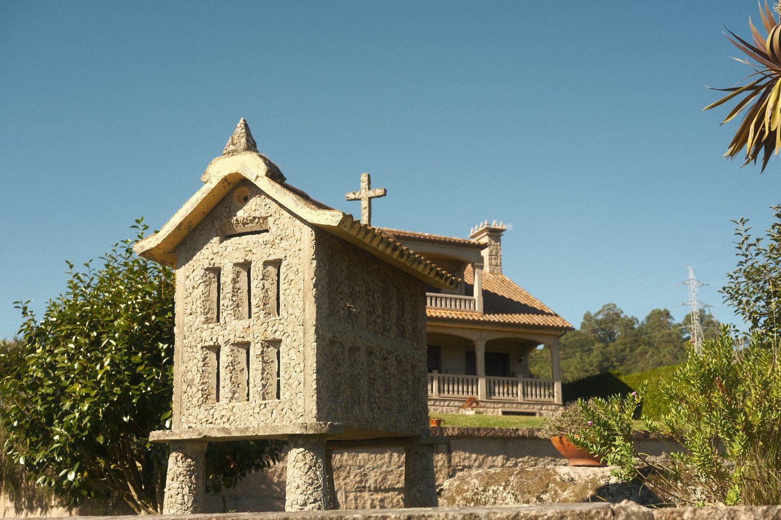 A rustic Galician hórreo stands under a clear sky next to a countryside house in Spain