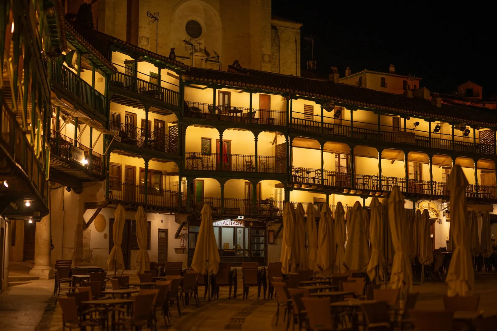 Illuminated arched balconies in Chinchón's Plaza Mayor with closed umbrellas and empty chairs creating a moody night scene