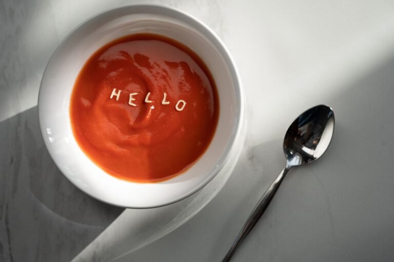 Tomato soup in a bowl with the word 'hello' formed by pasta letters, alongside a spoon