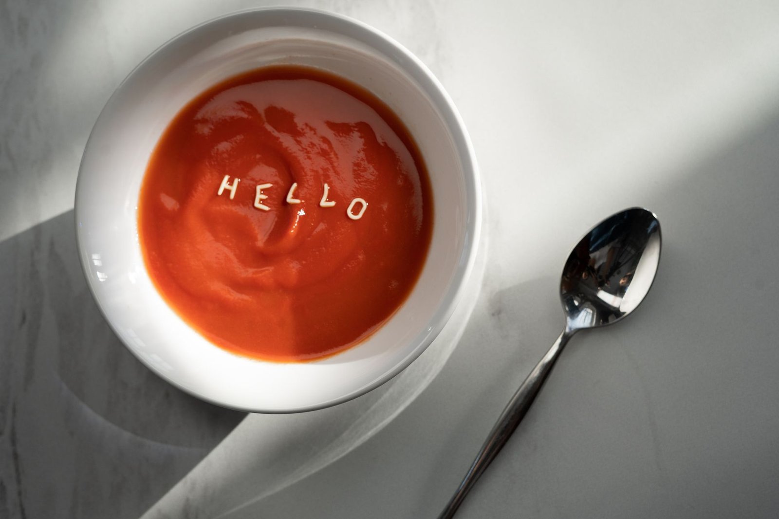 Tomato soup in a bowl with the word 'hello' formed by pasta letters, alongside a spoon