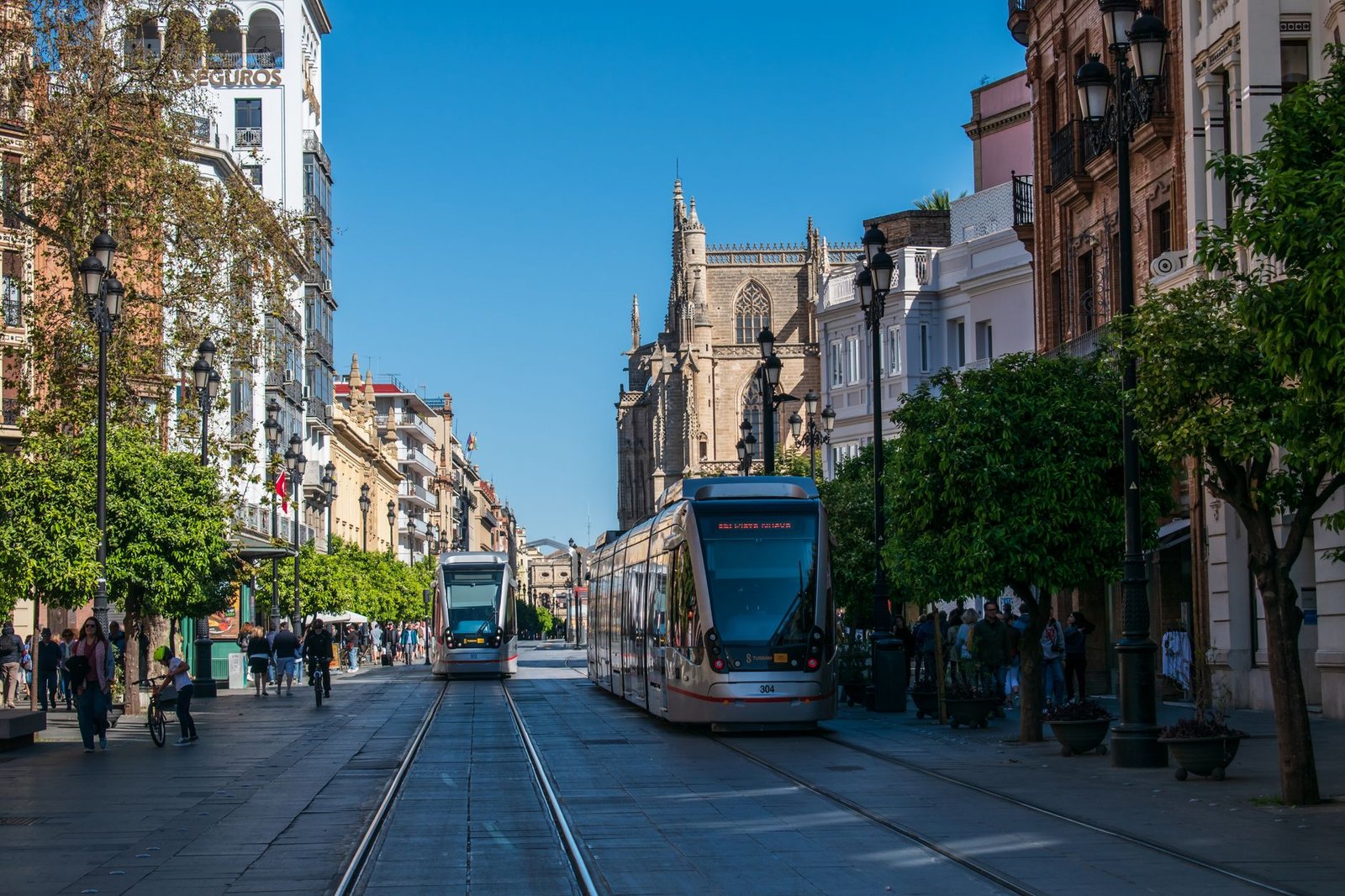 Trams traverse Avenida de la Constitución in Seville, showcasing urban architecture and street life