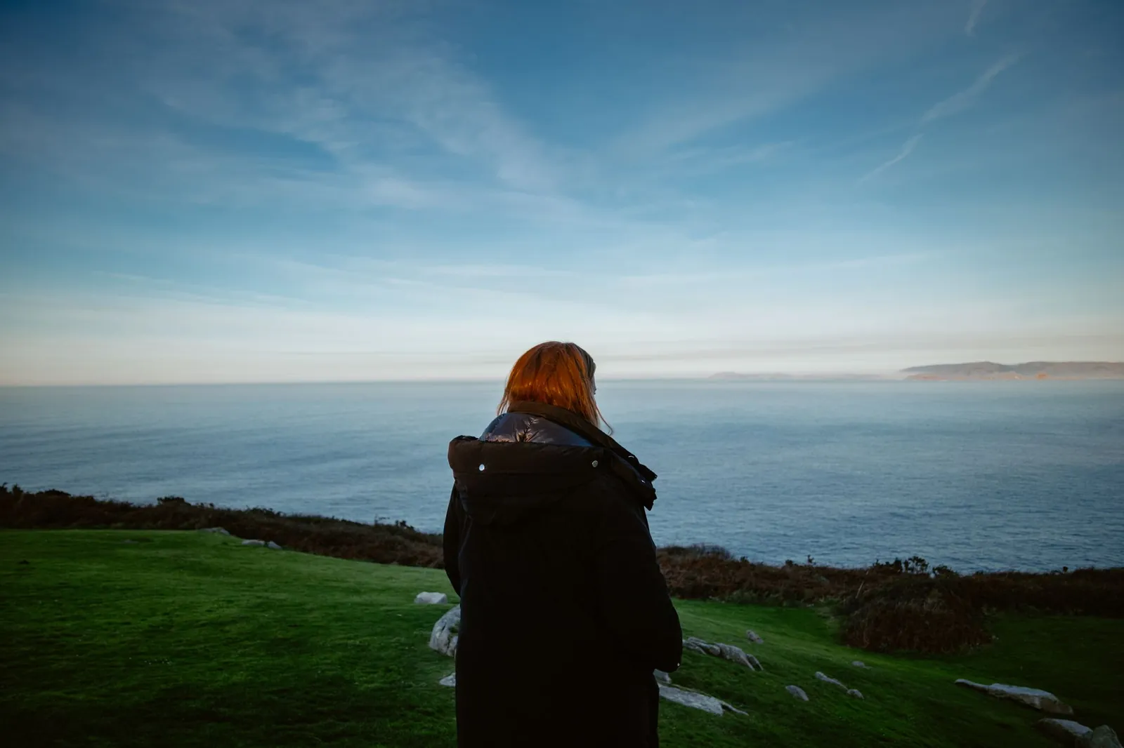 A redhead woman in a black jacket stands on a cliff overlooking the Atlantic Ocean in A Coruña, Spain