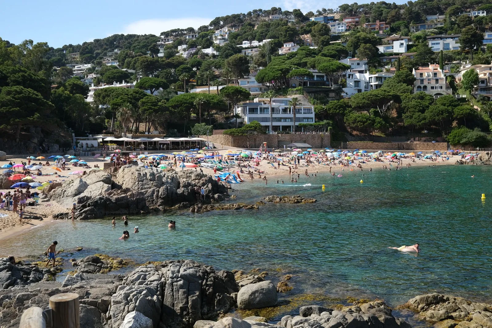 Crowded summer beach scene with turquoise waters and houses on Costa Brava